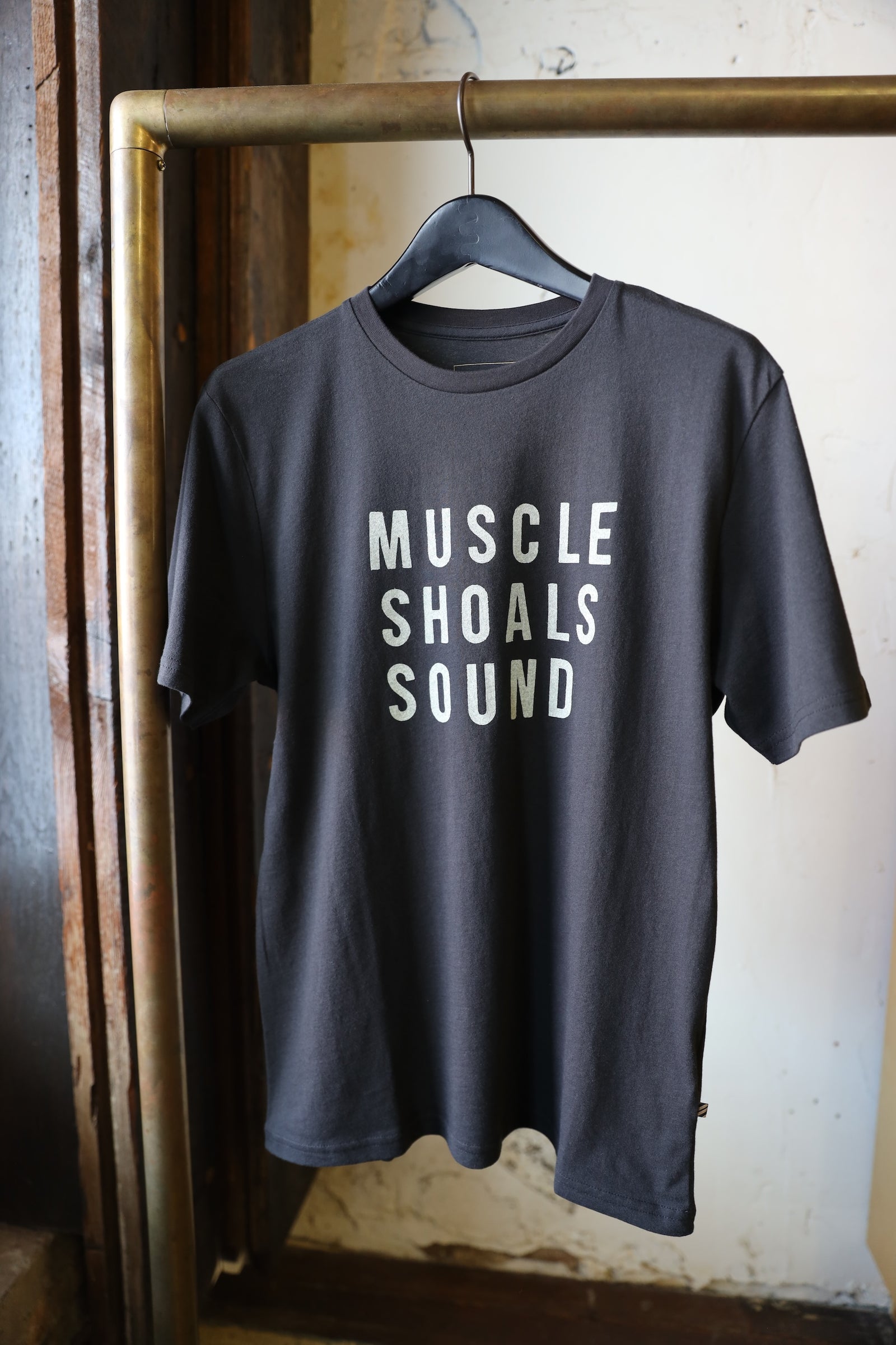 A black t-shirt hanging on a hanger from a brass clothing rack. The shirt features the text 'MUSCLE SHOALS SOUND' printed in a bold, white sans-serif font across the chest.