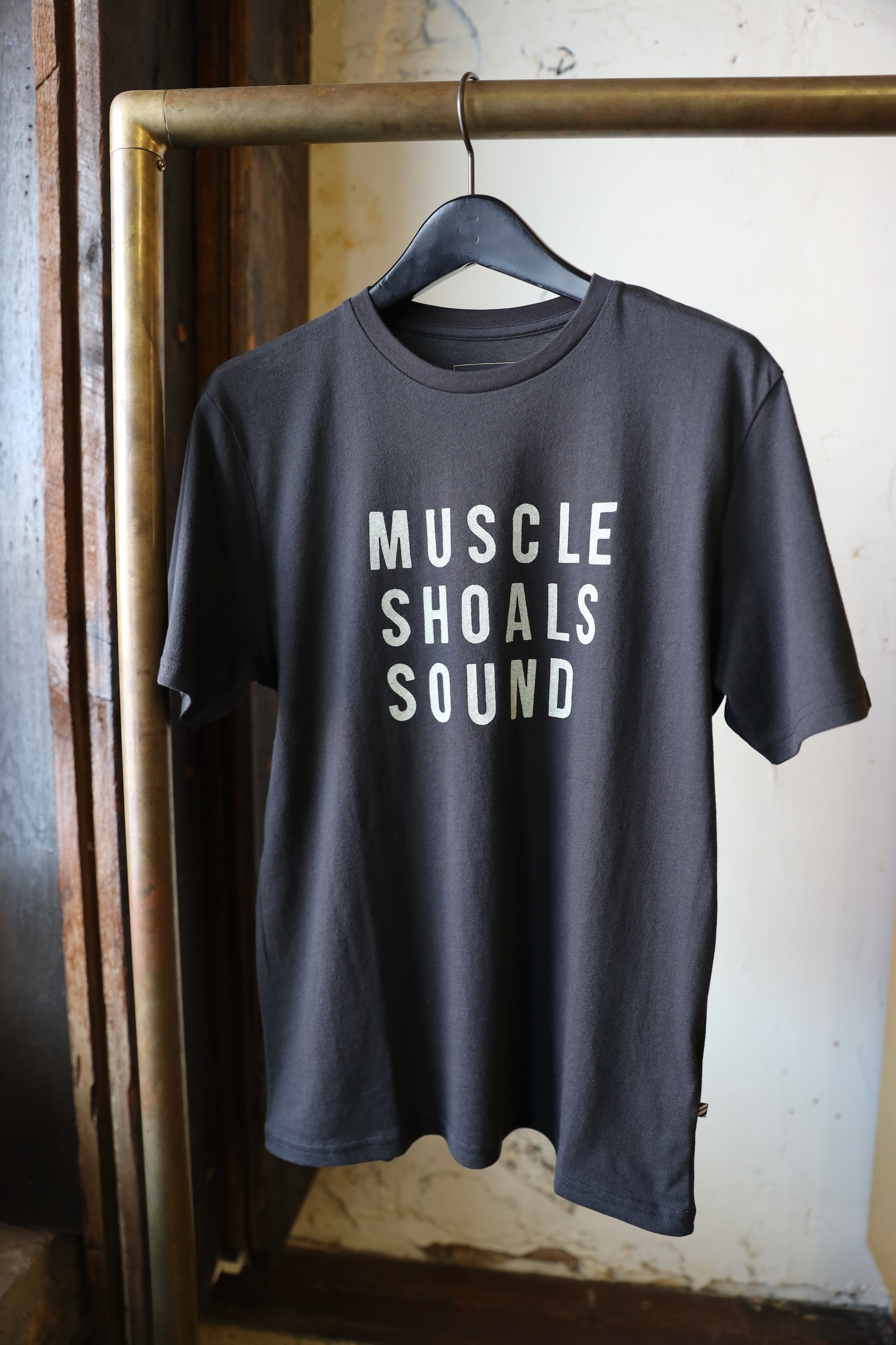 A dark charcoal gray t-shirt hanging on a black hanger from a brass clothing rack. The shirt features the text 'MUSCLE SHOALS SOUND' printed in a bold, light beige sans-serif font across the chest.
