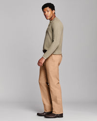 A man modeling a ribbed knit mock-neck sweater in a light olive-beige color, paired with tan trousers and dark brown leather shoes, photographed against a solid light gray studio background.