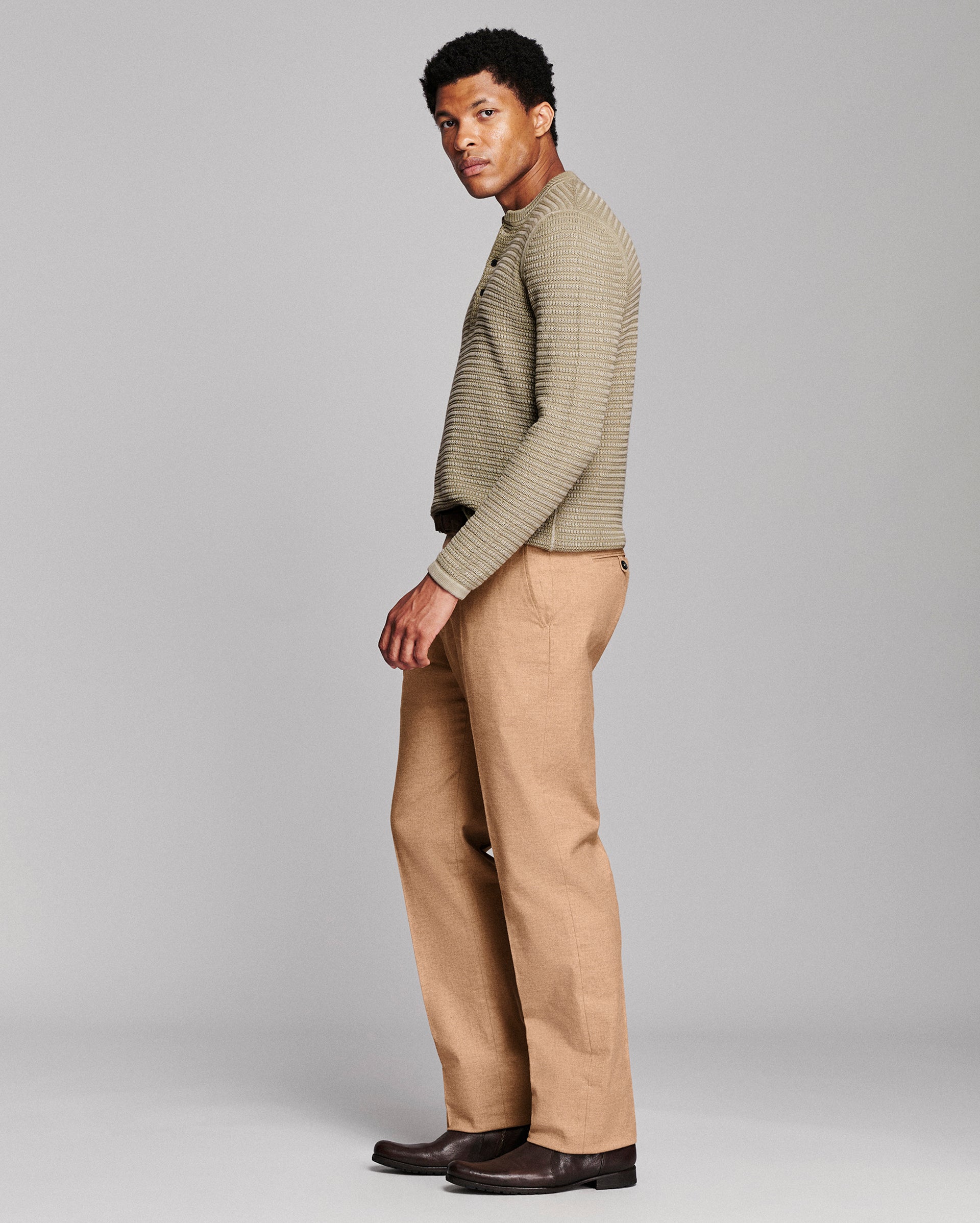A man modeling a ribbed knit mock-neck sweater in a light olive-beige color, paired with tan trousers and dark brown leather shoes, photographed against a solid light gray studio background.