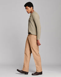 A man wearing a ribbed knit long-sleeve sweater in a light olive-beige color and tan trousers, shown in profile against a neutral gray studio background.