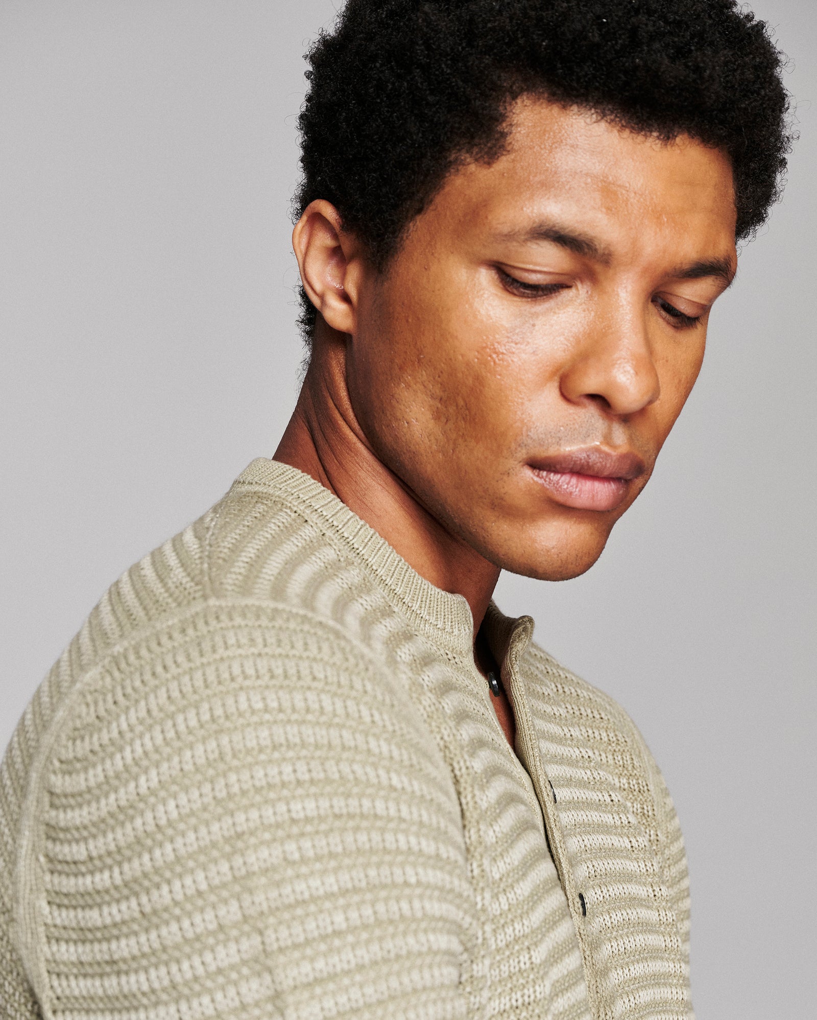 A close-up side profile of a man wearing a textured, ribbed knit half-zip sweater in a light moss green color, set against a solid gray studio background.