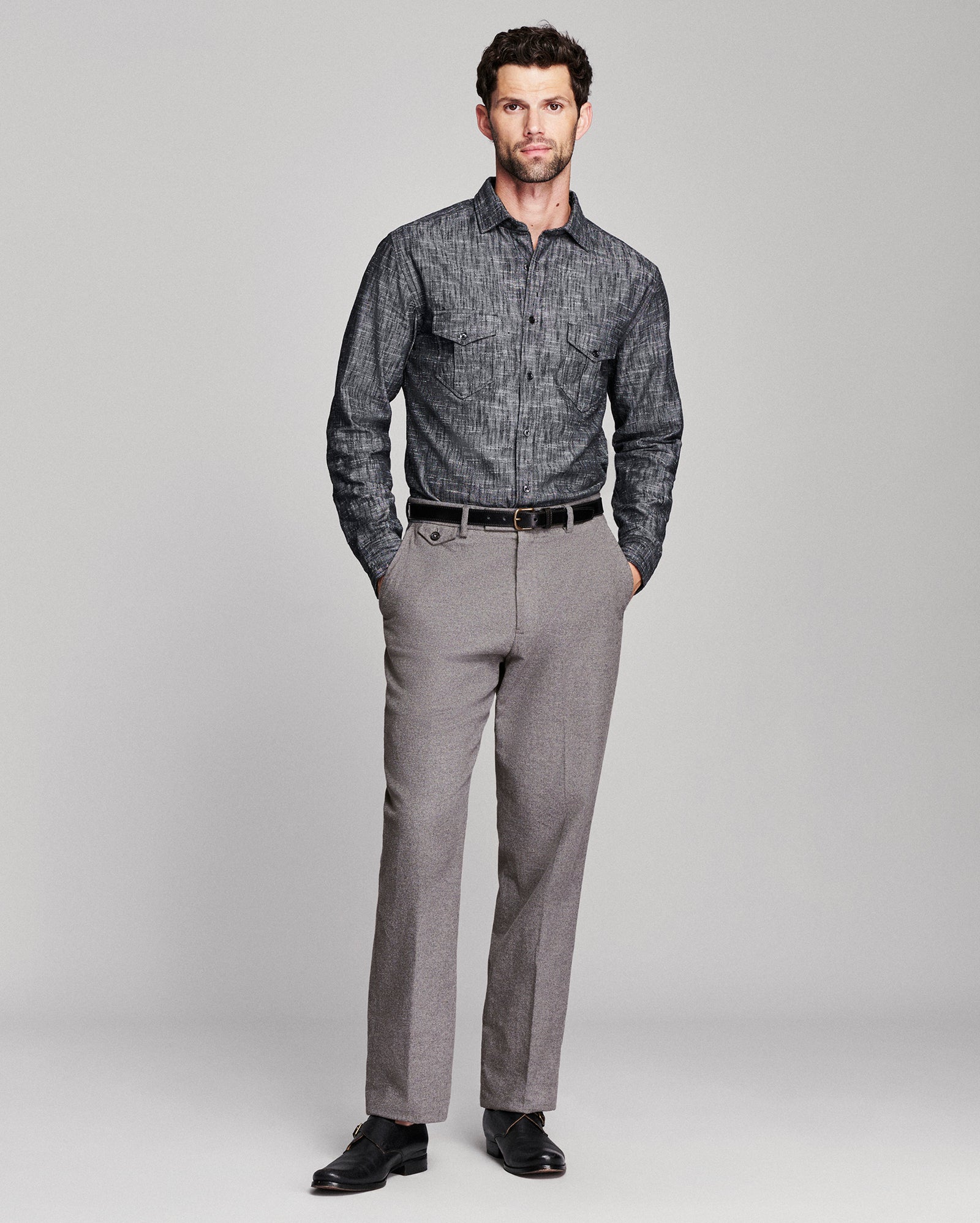 A man modeling a charcoal gray patterned long-sleeve button-down shirt paired with gray trousers, a black belt, and black dress shoes against a light gray studio background.
