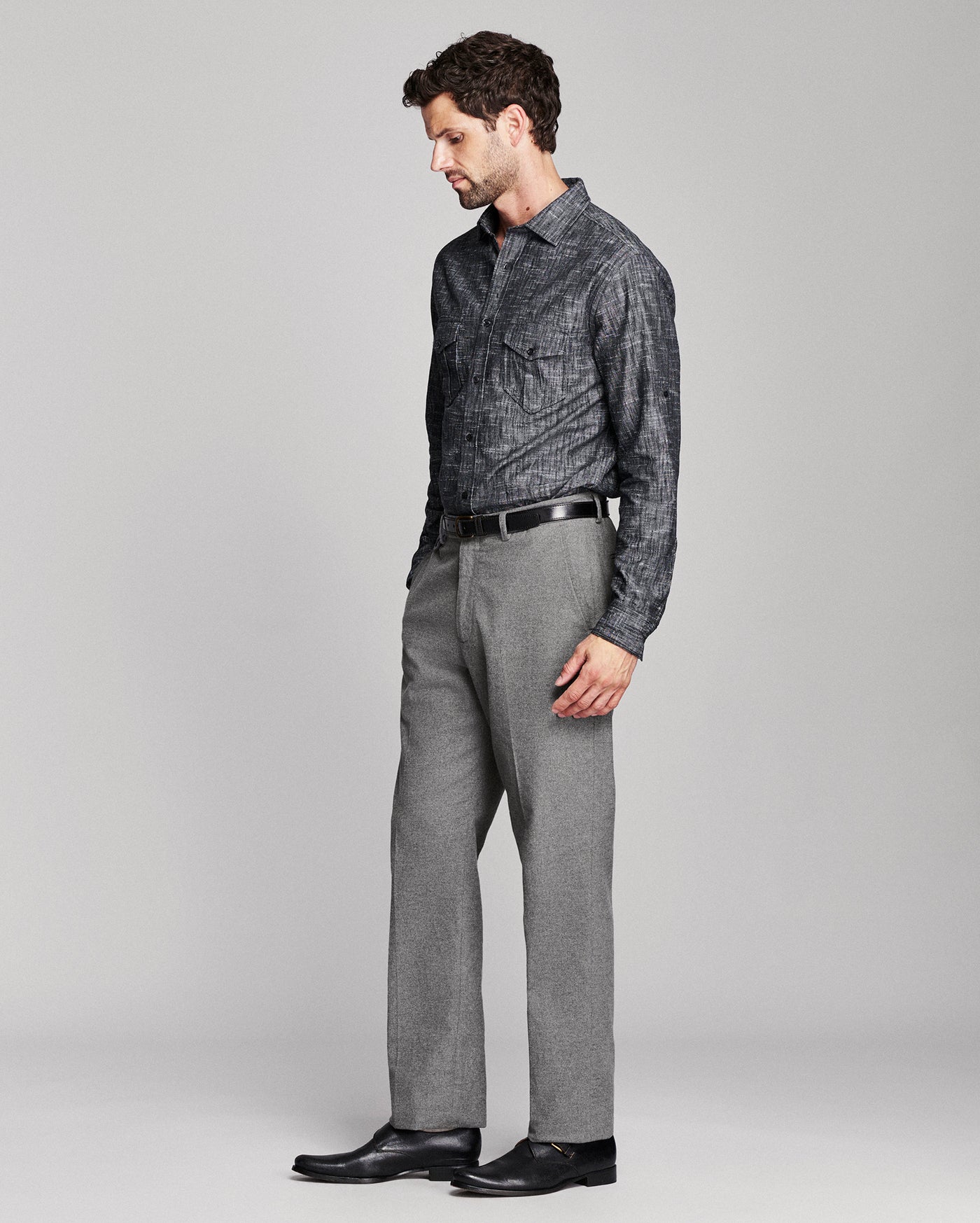 A man wearing a dark gray patterned button-down shirt, gray trousers, a black belt, and black dress shoes, standing in profile against a light gray studio background.
