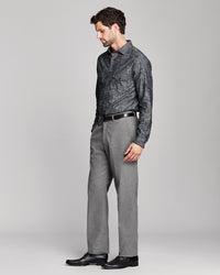 A man wearing a dark gray patterned button-down shirt, gray trousers, a black belt, and black dress shoes, standing in profile against a light gray studio background.