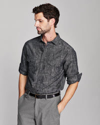 A man wearing a dark gray textured short-sleeve button-up shirt with rolled-up sleeves and gray trousers, photographed against a solid light gray studio background.