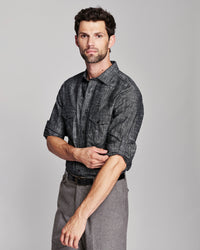 A man wearing a dark gray and black textured long-sleeve button-down shirt with rolled-up sleeves, paired with gray trousers, photographed against a solid light gray background.