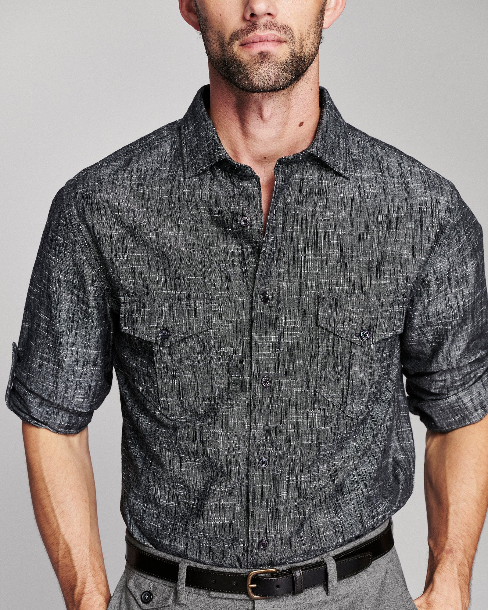 A man wearing a dark gray textured short-sleeve button-up shirt with two chest pockets, paired with gray trousers and a black belt, against a light gray studio background.