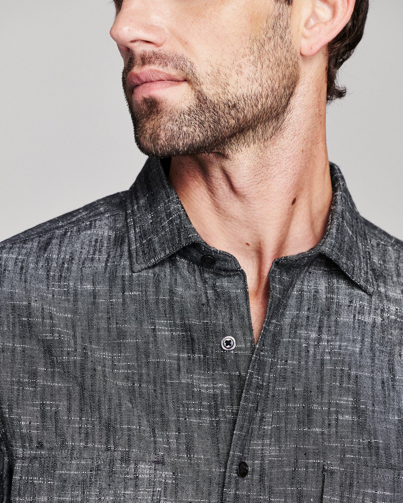 Close-up of a man wearing a dark gray textured button-down shirt, showing the collar, chest area, and part of his face against a light gray background.