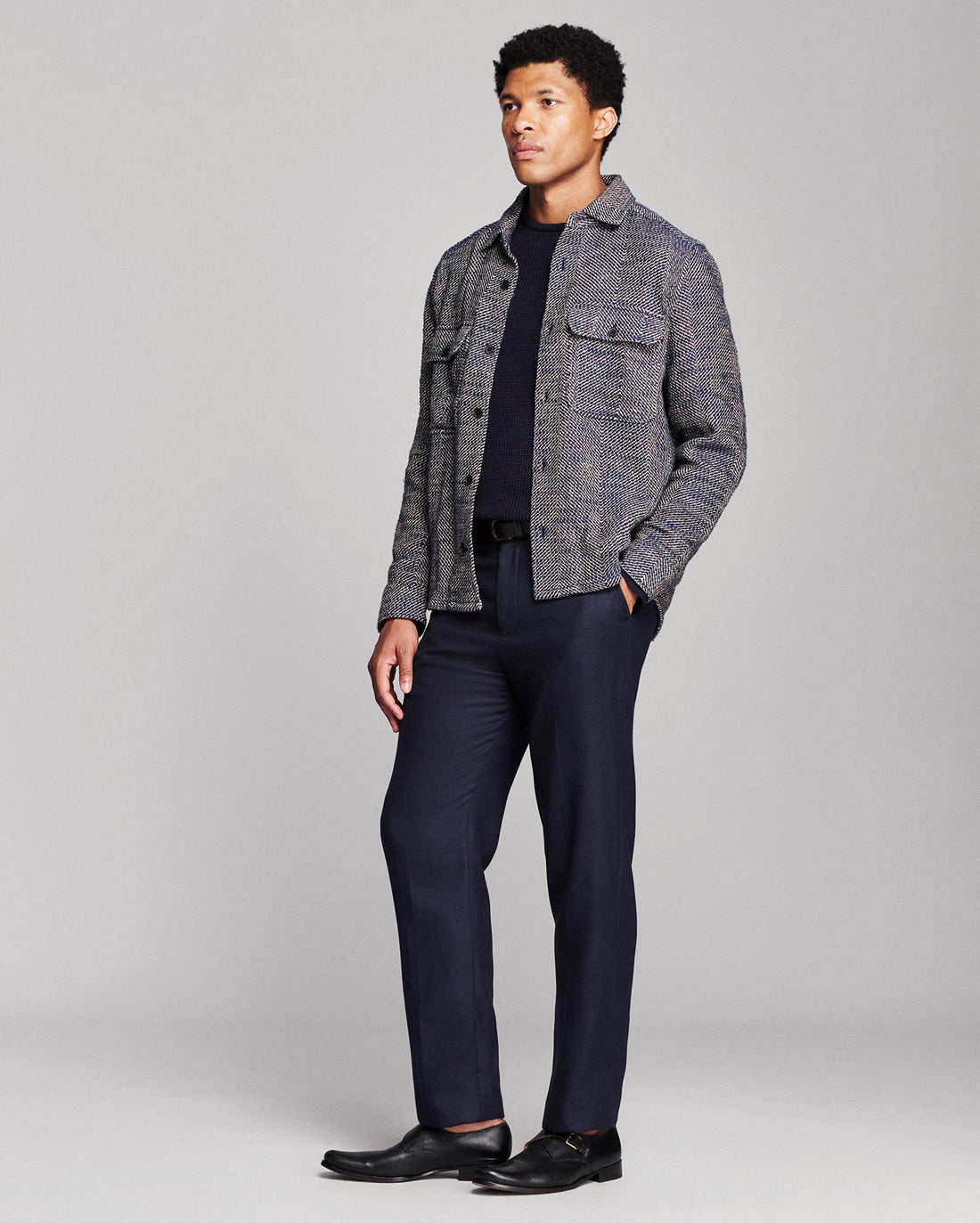 A man modeling a gray and navy tweed button-up shirt jacket over a dark navy t-shirt, paired with navy trousers and black leather dress shoes, against a light gray studio background.