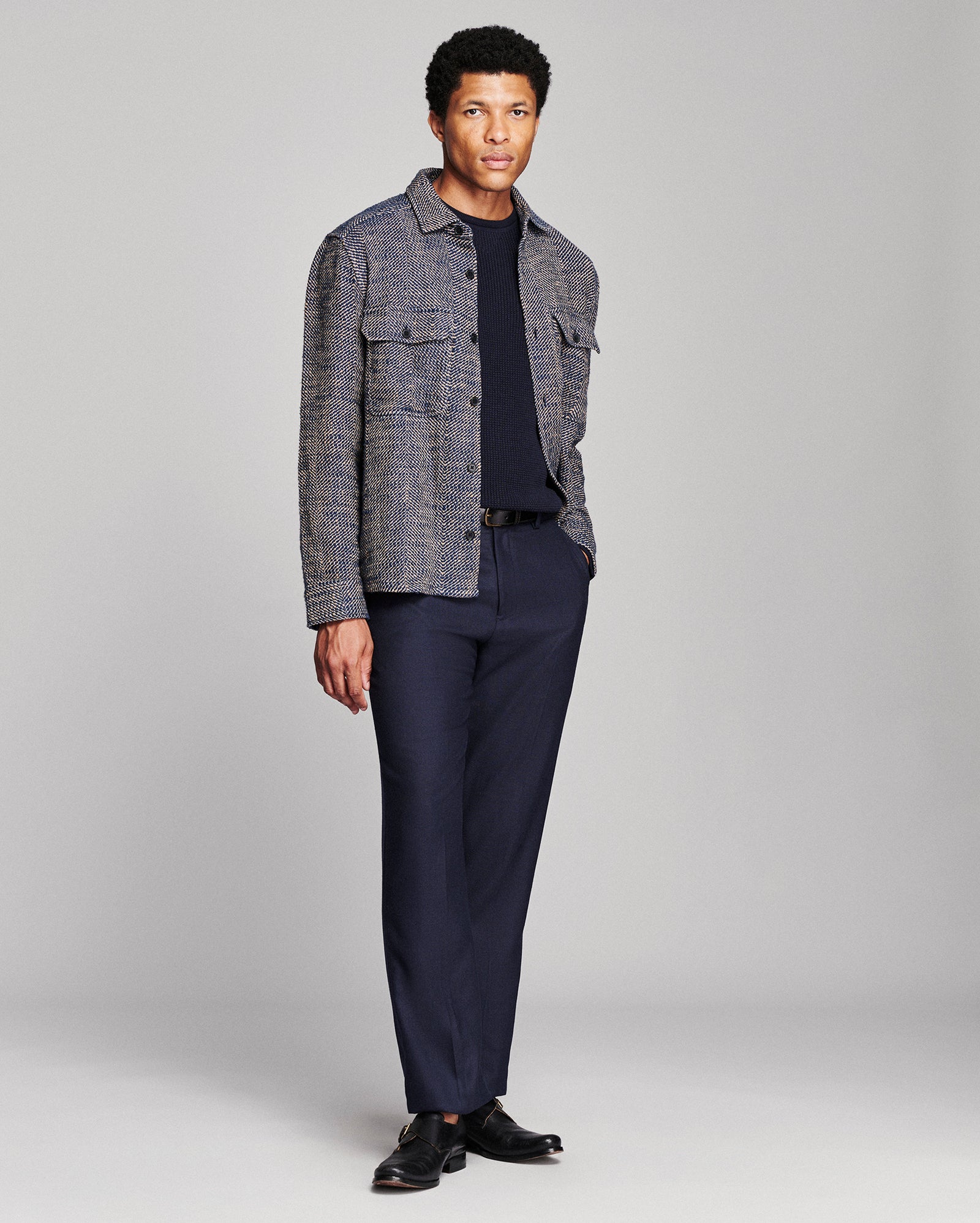 A man modeling a gray textured overshirt jacket over a dark navy crewneck shirt and matching navy trousers, paired with black dress shoes, against a light gray studio background.