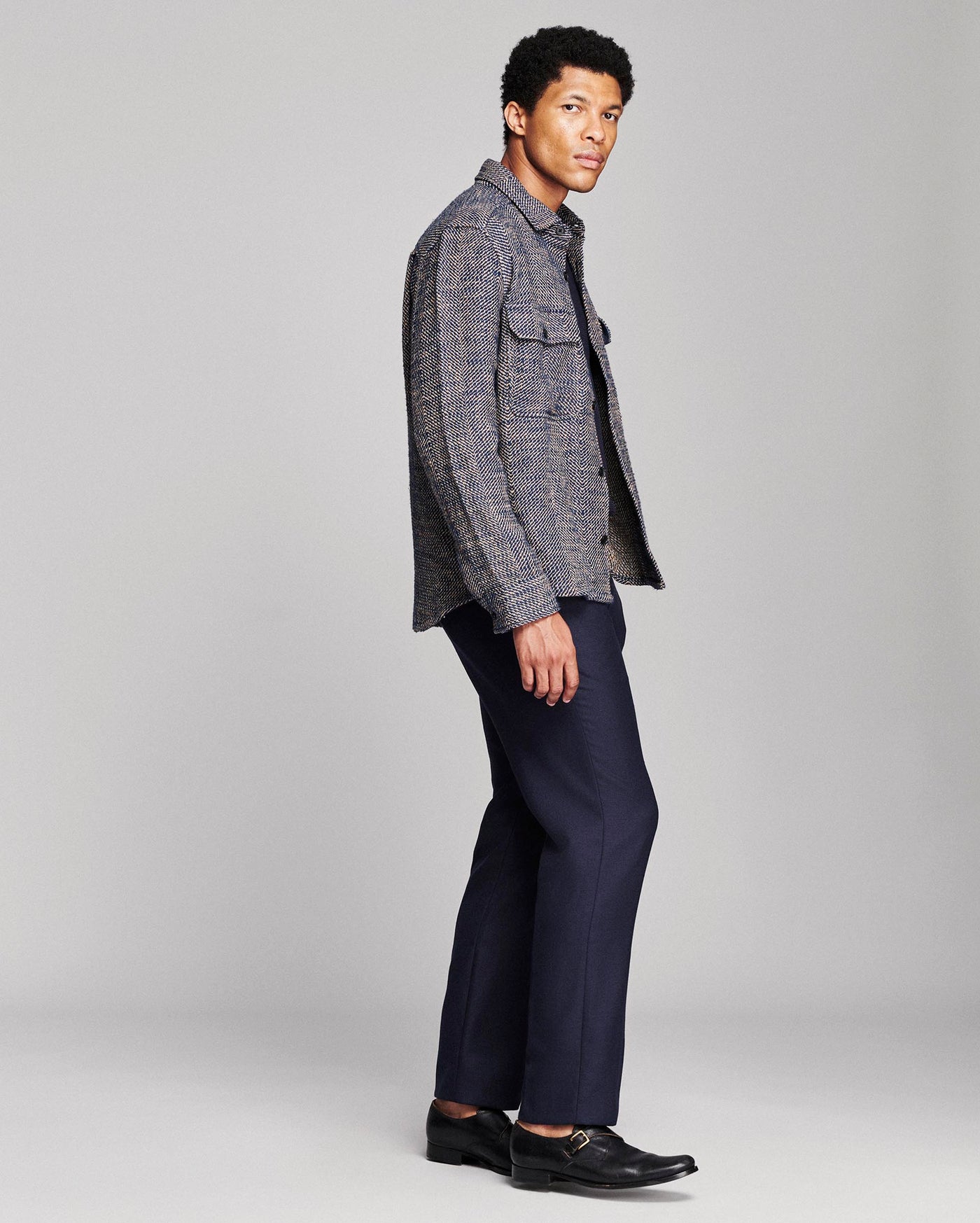 A man in a gray textured button-up shirt jacket and navy blue trousers, standing in profile against a light gray studio background.