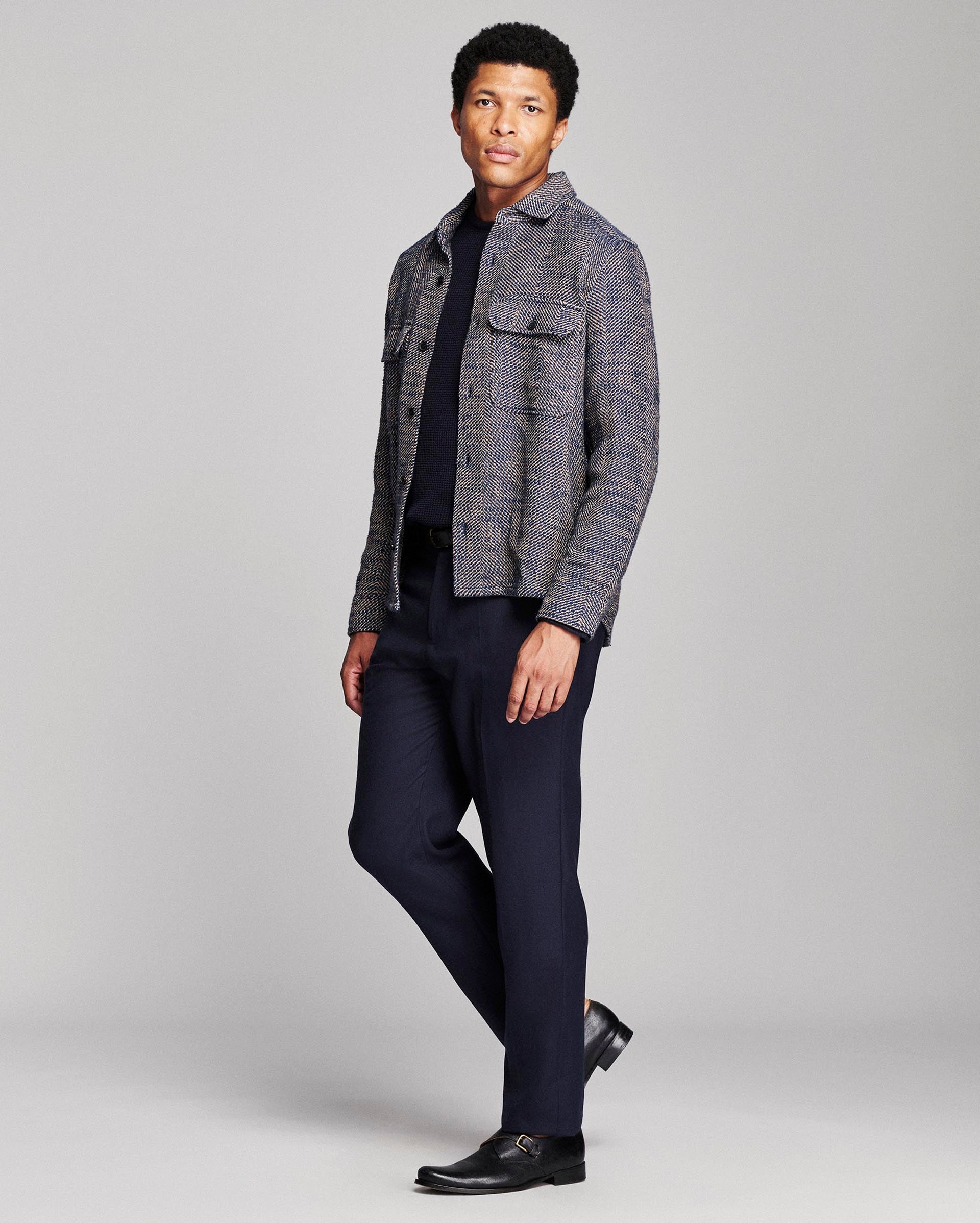 A man modeling a gray and black textured tweed jacket over a dark navy shirt and navy trousers, paired with black dress shoes, against a light gray studio background.