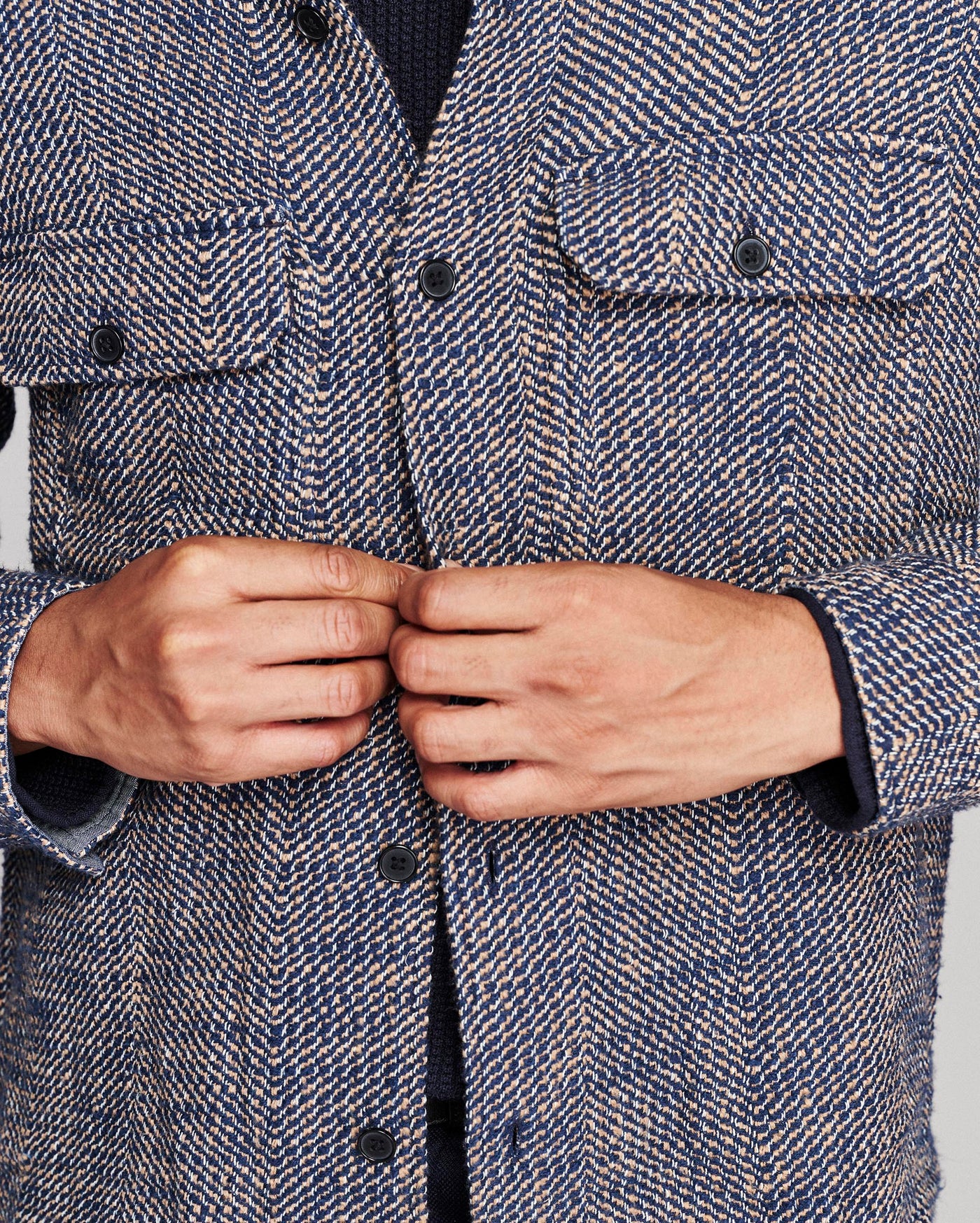 Close-up of a person wearing a textured herringbone tweed jacket, buttoning the front. The jacket features a blue, gray, and white woven pattern with dark buttons and flap chest pockets.