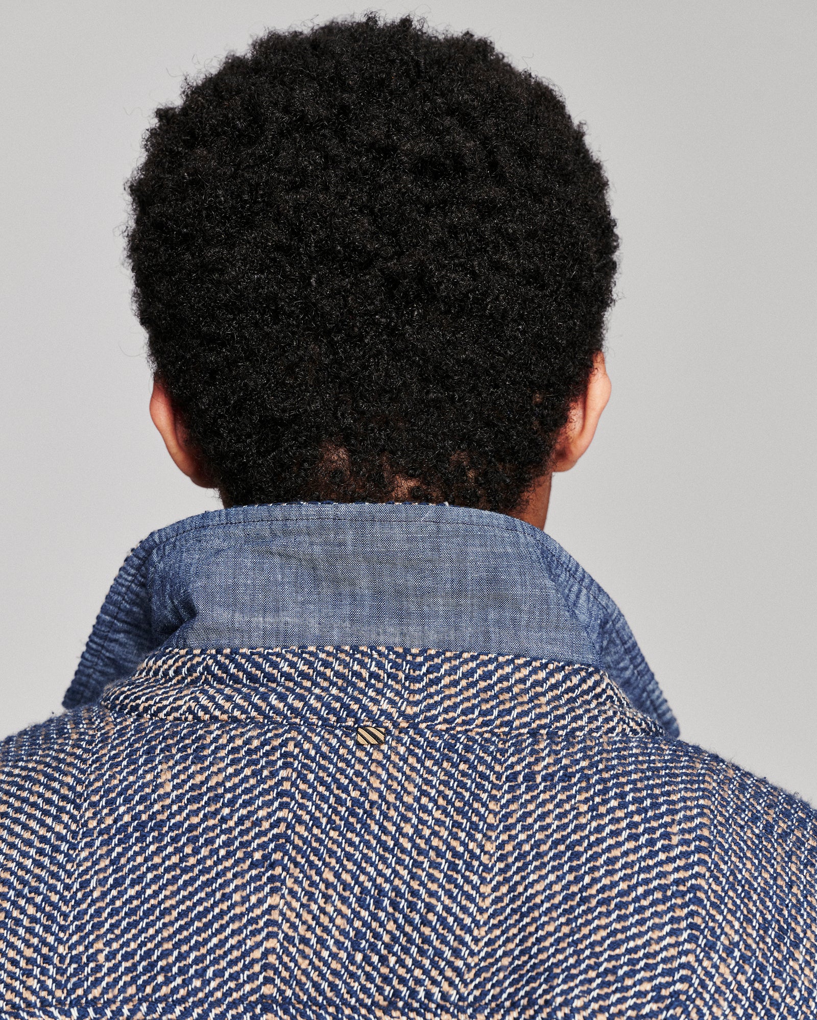 Back view of a person with short, curly black hair wearing a textured blue and beige herringbone jacket against a solid gray background.
