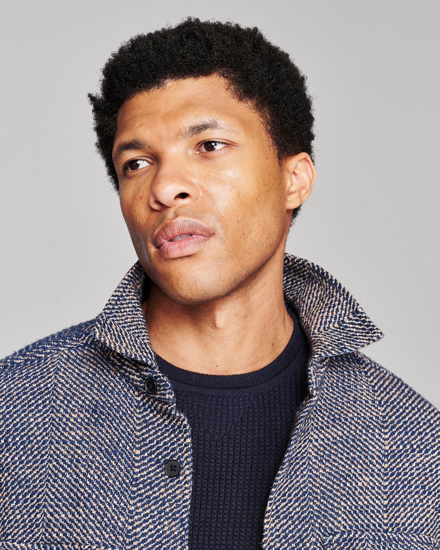 A close-up portrait of a man with short black curly hair, wearing a textured blue and white herringbone jacket over a dark navy knit shirt, set against a solid light gray background.
