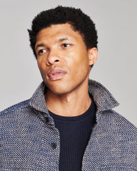 A close-up portrait of a man with short black curly hair, wearing a textured blue and white herringbone jacket over a dark navy knit shirt, set against a solid light gray background.