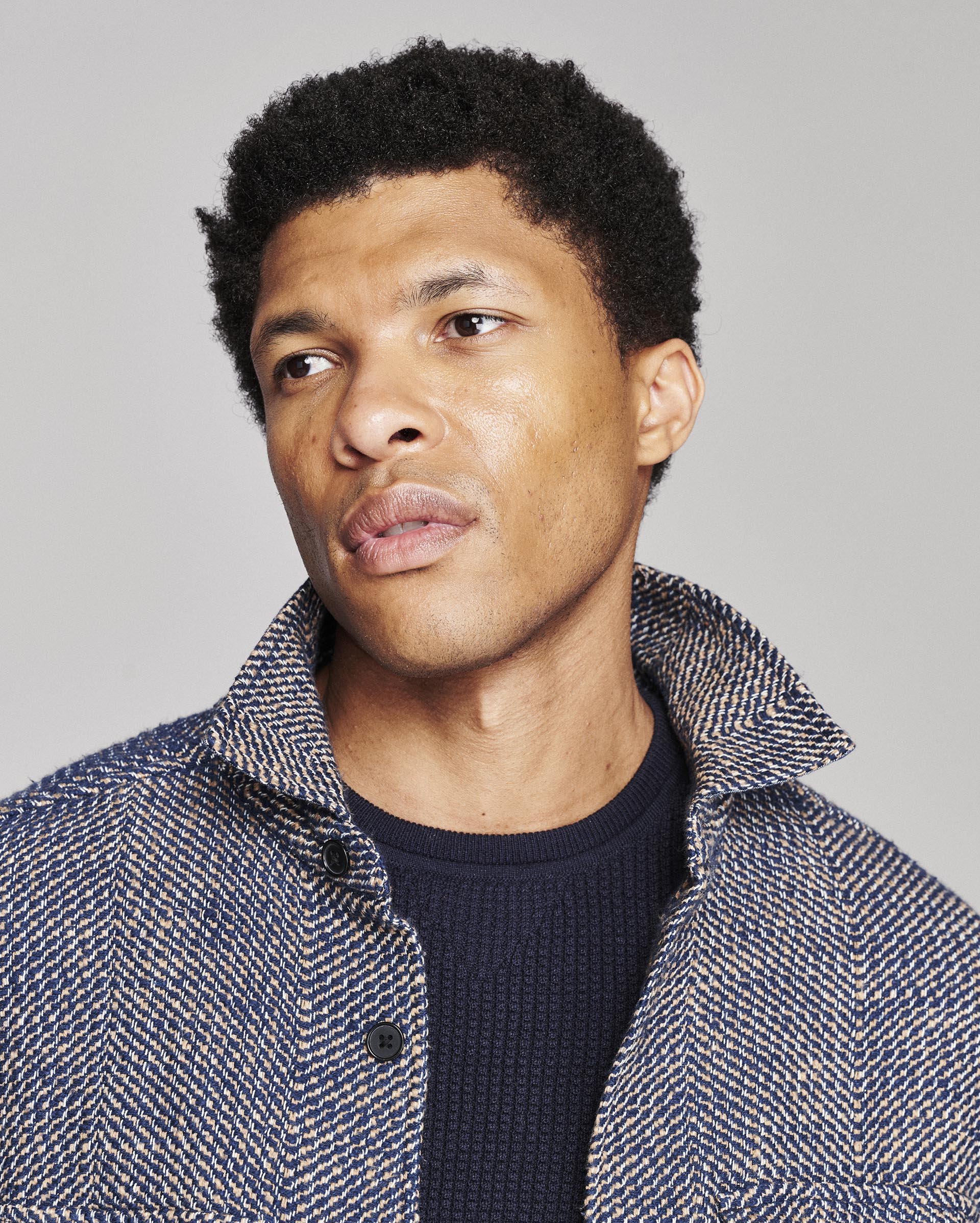A close-up portrait of a man with short black curly hair, wearing a textured blue and white herringbone jacket over a dark navy knit shirt, set against a solid light gray background.