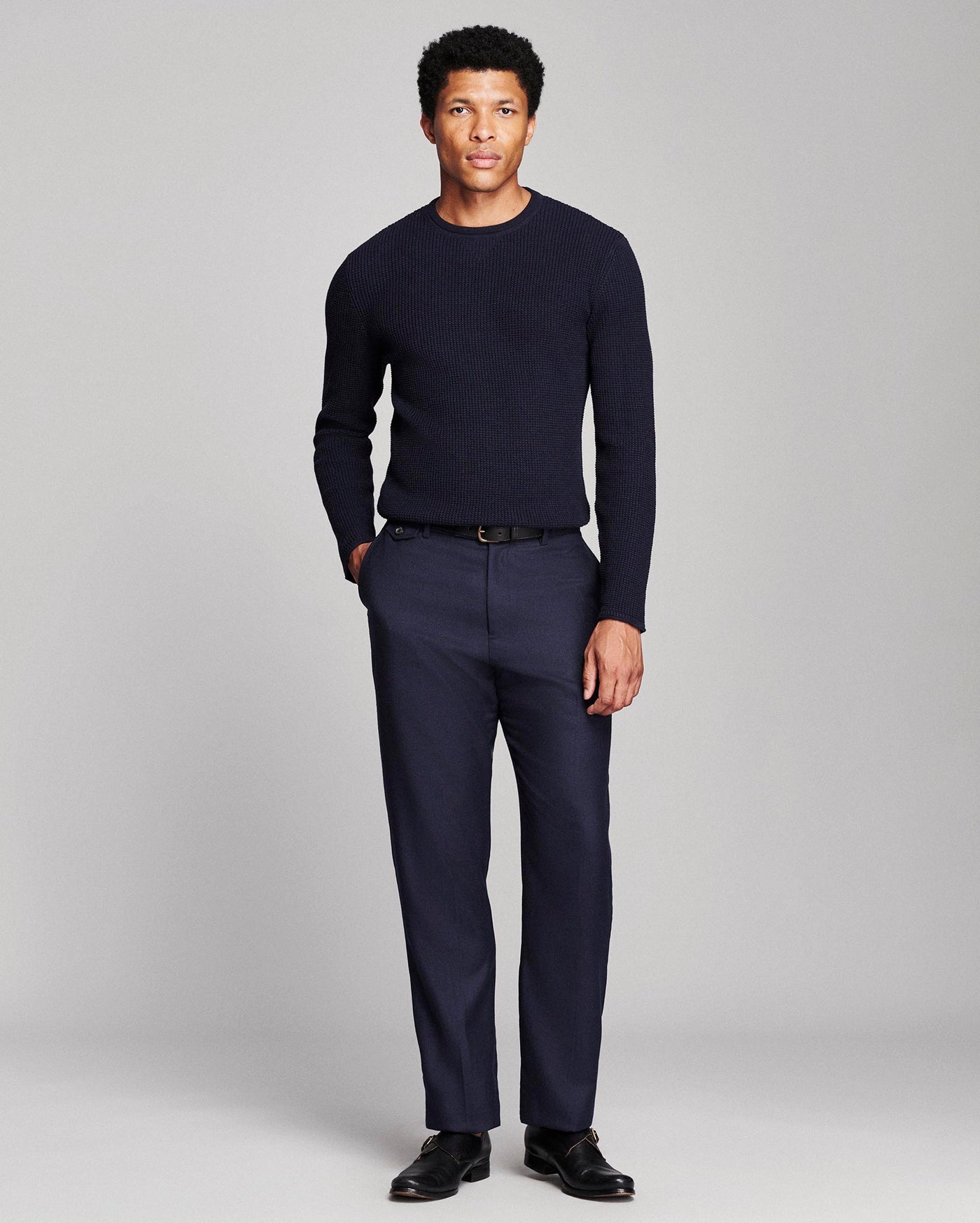 A man modeling a dark navy blue long-sleeve knit sweater and matching navy blue trousers with a black belt and black dress shoes, photographed against a neutral light gray studio background.