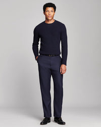 A man modeling a dark navy blue long-sleeve knit sweater and matching navy blue trousers with a black belt and black dress shoes, photographed against a neutral light gray studio background.