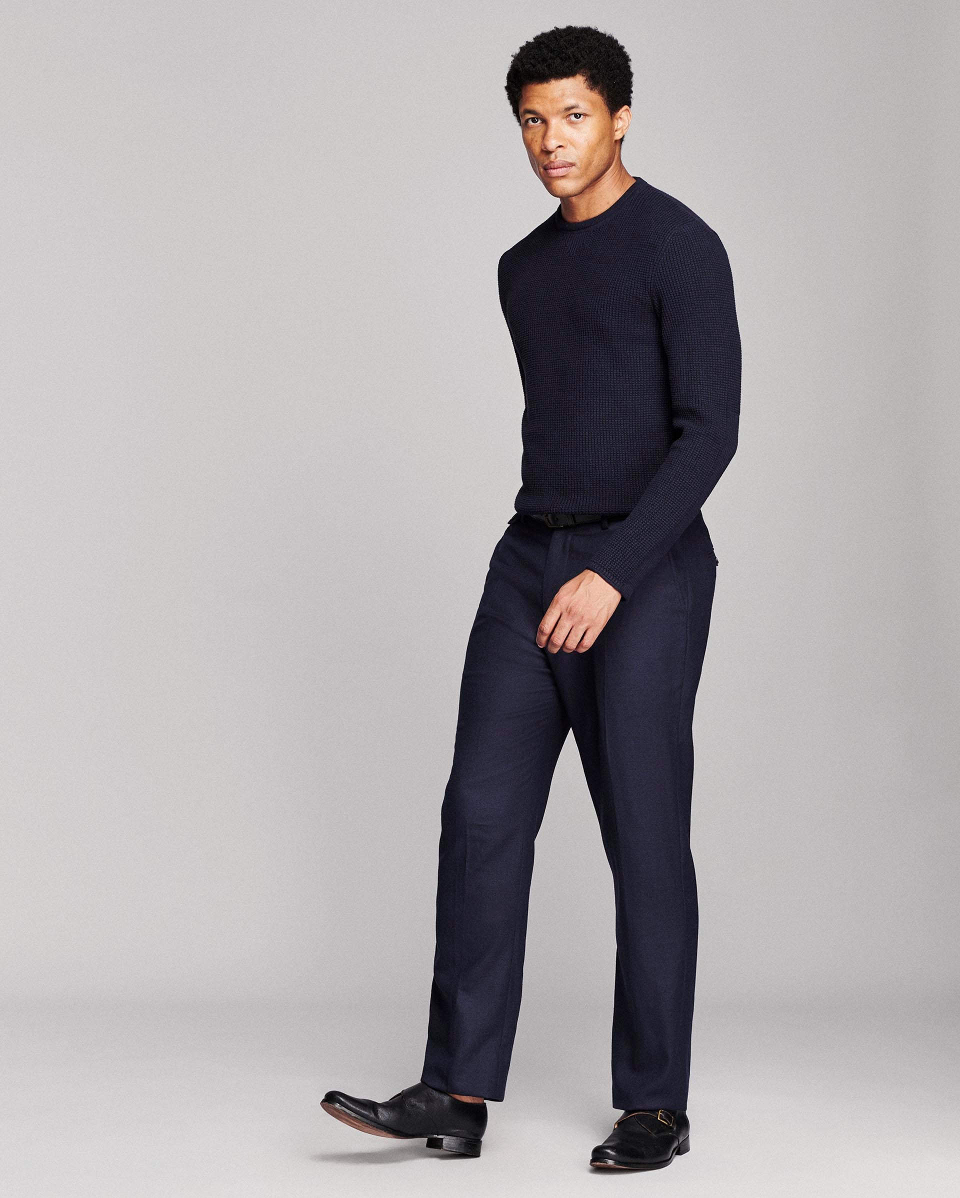 A man modeling a dark navy blue knit sweater and matching navy trousers with black dress shoes, standing against a light gray studio background.