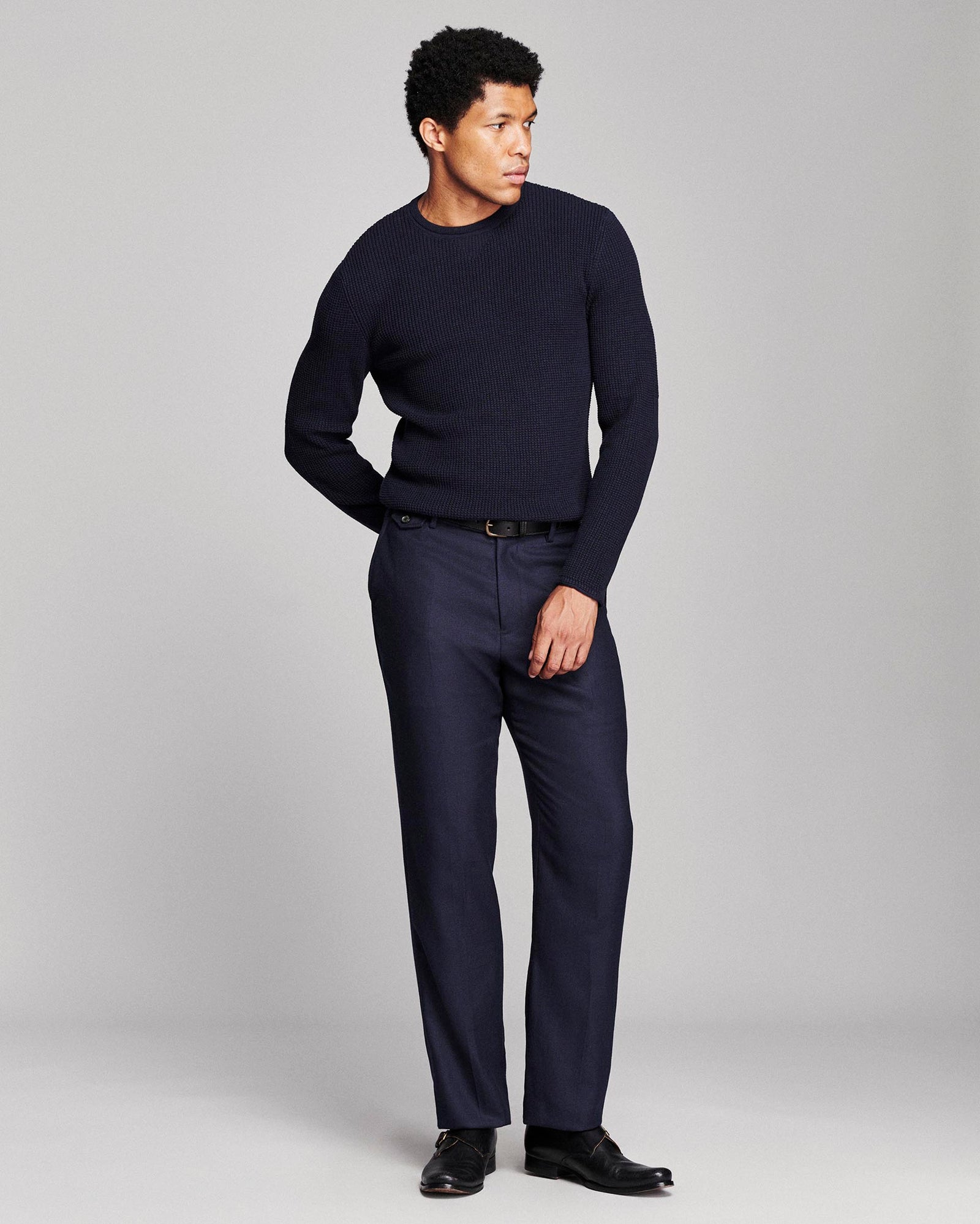 A man modeling a dark navy blue crewneck sweater and matching navy trousers with black dress shoes, standing against a light gray studio background.