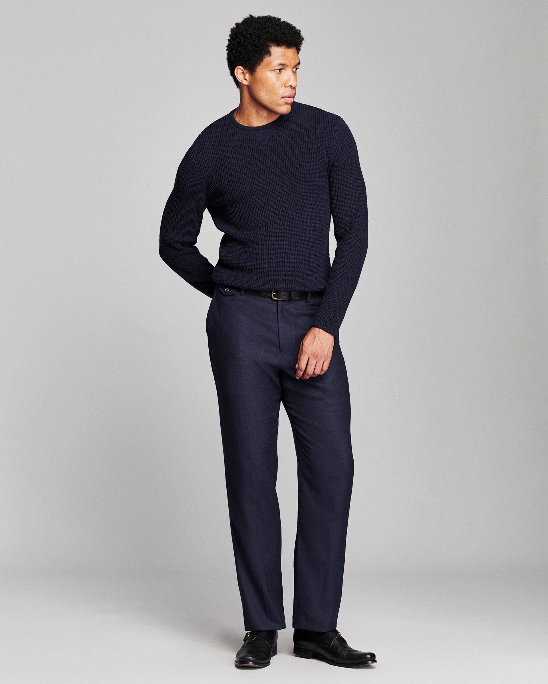 A man modeling a dark navy blue crewneck sweater and matching navy trousers with black dress shoes, standing against a light gray studio background.