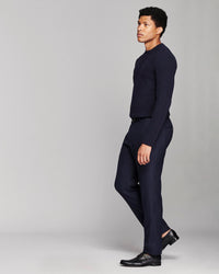 A man in a dark navy blue knit sweater and matching navy trousers, styled with black leather dress shoes, standing in profile against a light gray studio background.