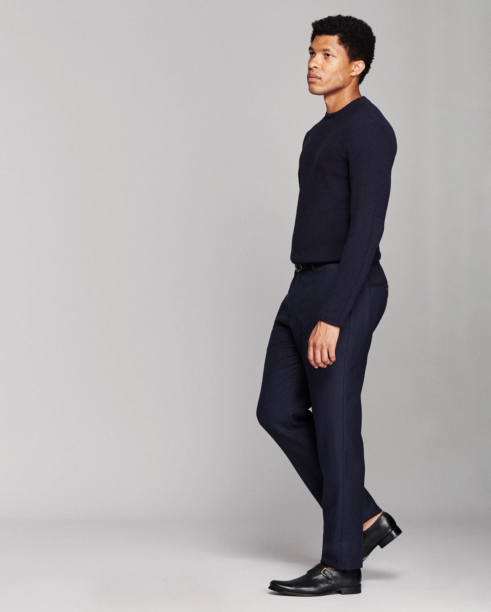 A man in a dark navy blue knit sweater and matching navy trousers, styled with black leather dress shoes, standing in profile against a light gray studio background.