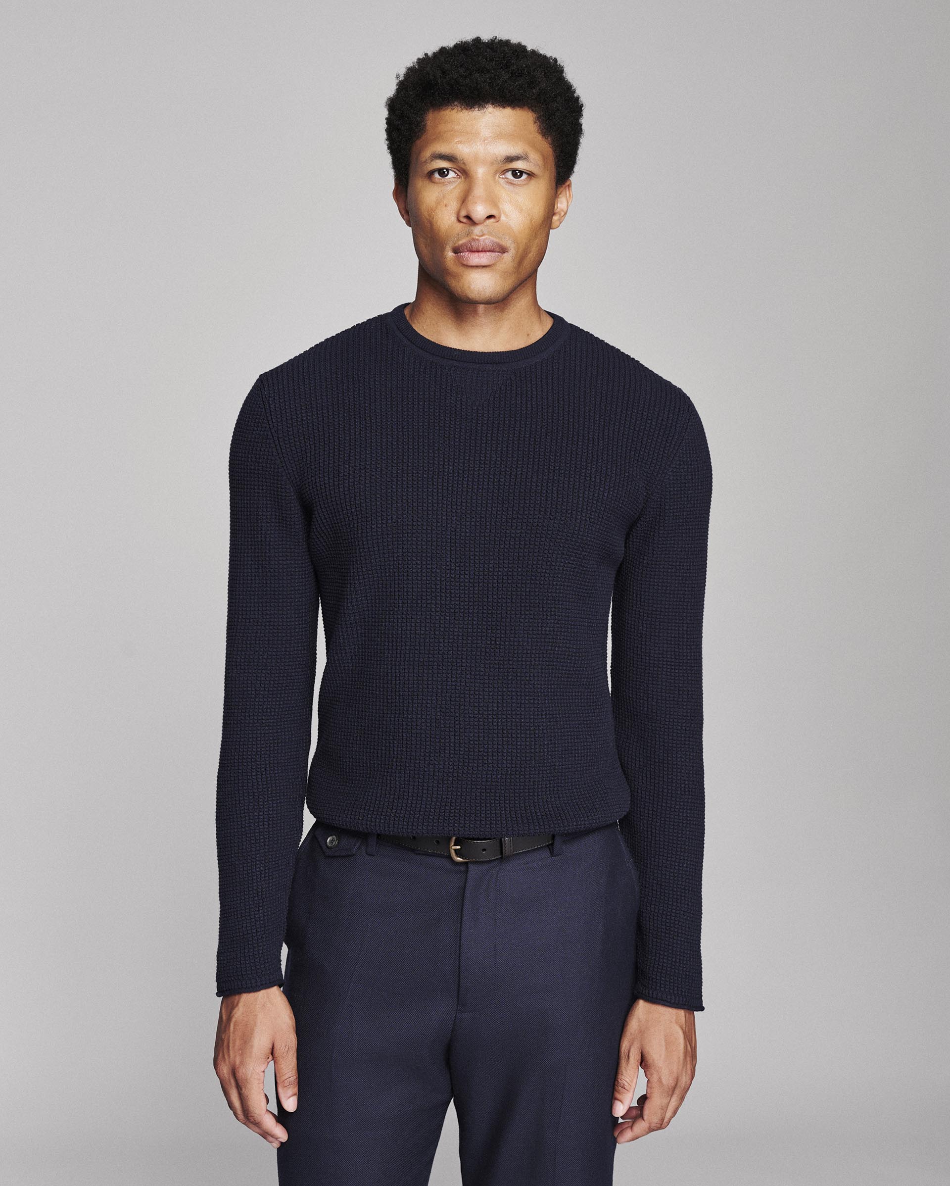 A man wearing a dark navy ribbed knit crewneck sweater and matching navy trousers, standing against a neutral gray studio background.