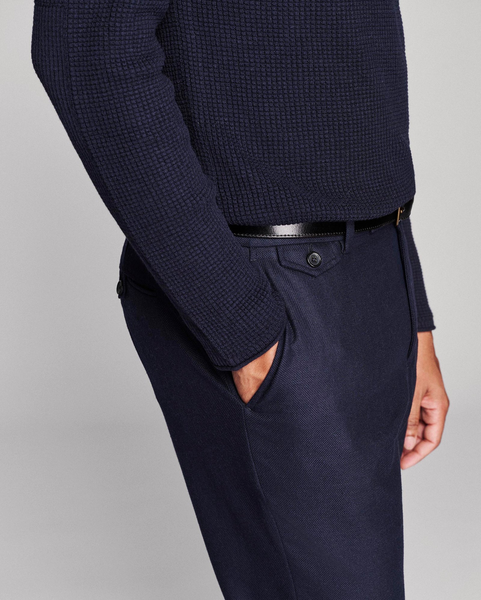 A close-up of a person wearing navy blue textured knit sweater and matching navy trousers with a black belt, shown against a light gray studio background.