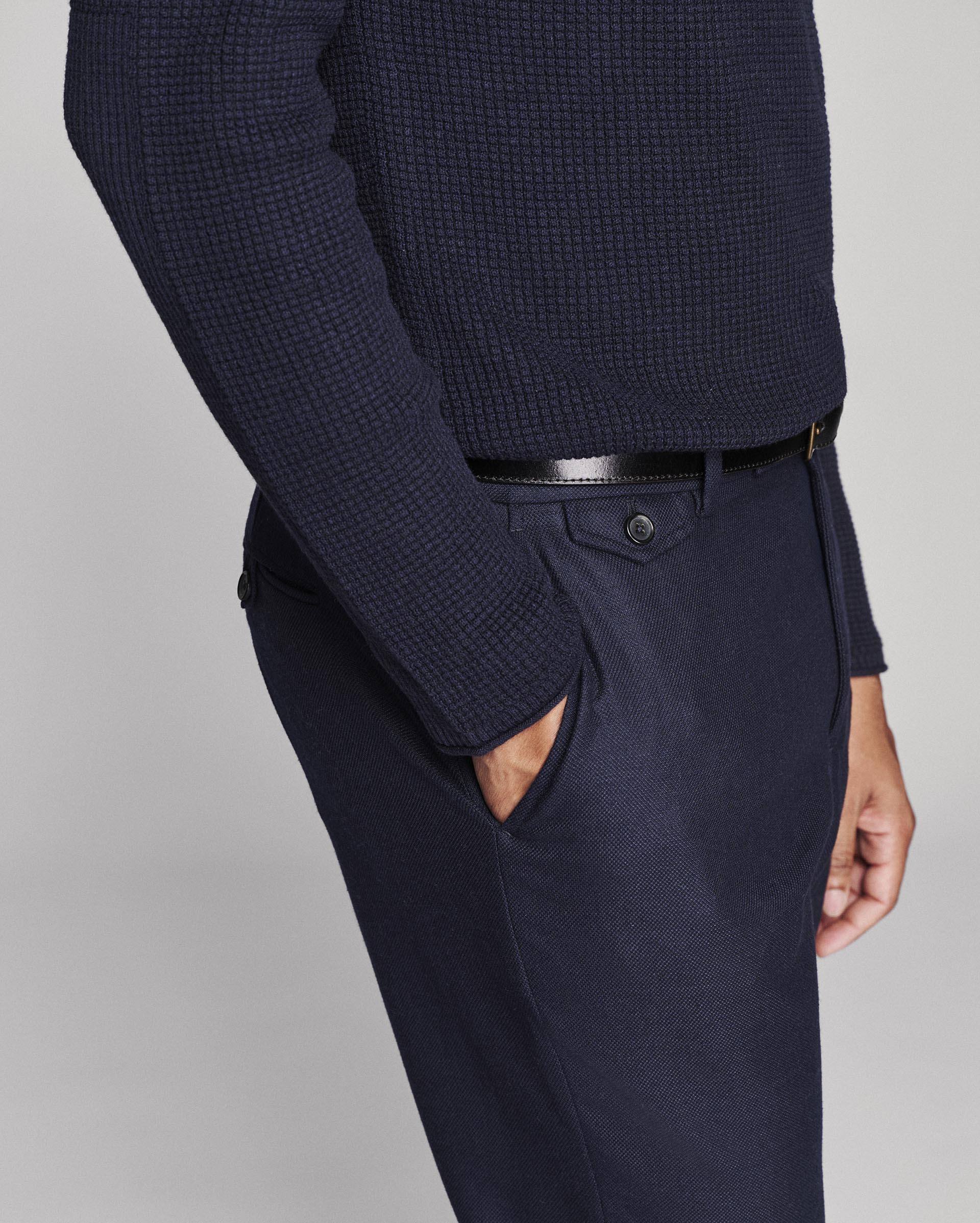 A close-up of a person wearing navy blue textured knit sweater and matching navy trousers with a black belt, shown against a light gray studio background.