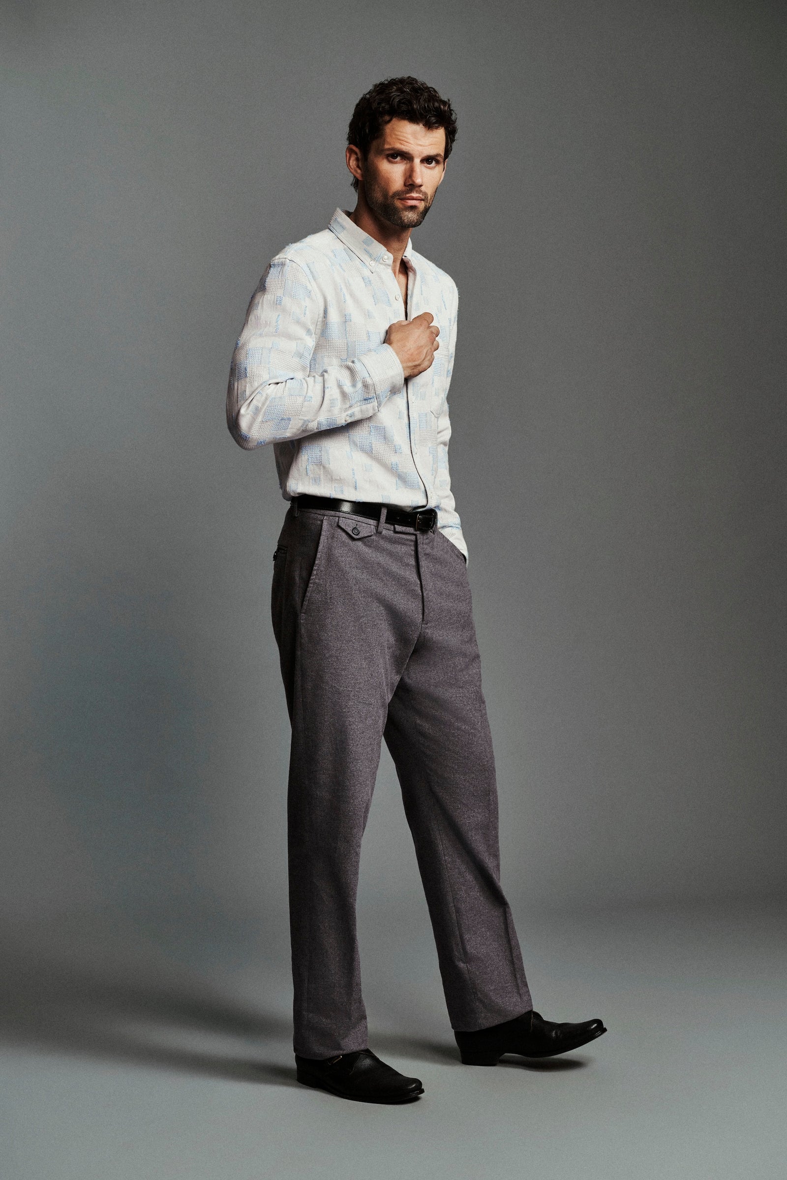 A man modeling a white long-sleeve button-down shirt and gray trousers against a solid gray studio background.