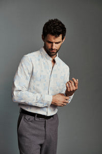 A man wearing a light blue and white patterned button-down shirt and gray trousers, adjusting his shirt cuff against a solid gray studio background.
