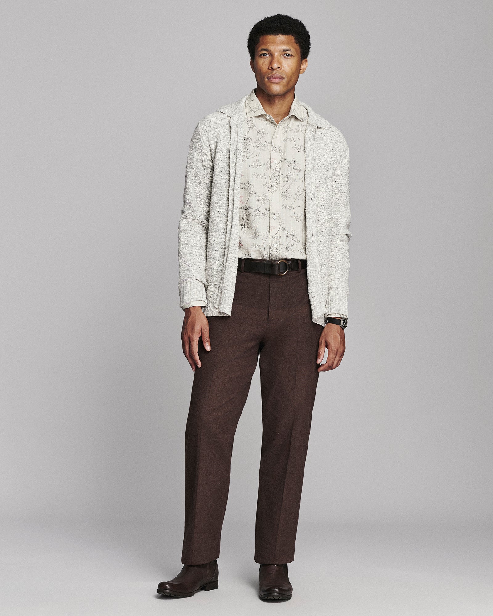 A man modeling a light beige patterned cardigan over a matching shirt, paired with dark brown trousers and dark brown shoes, against a solid gray studio background.