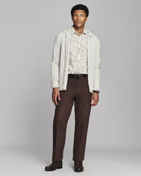 A man modeling a light beige patterned cardigan over a matching shirt, paired with dark brown trousers and dark brown shoes, against a solid gray studio background.