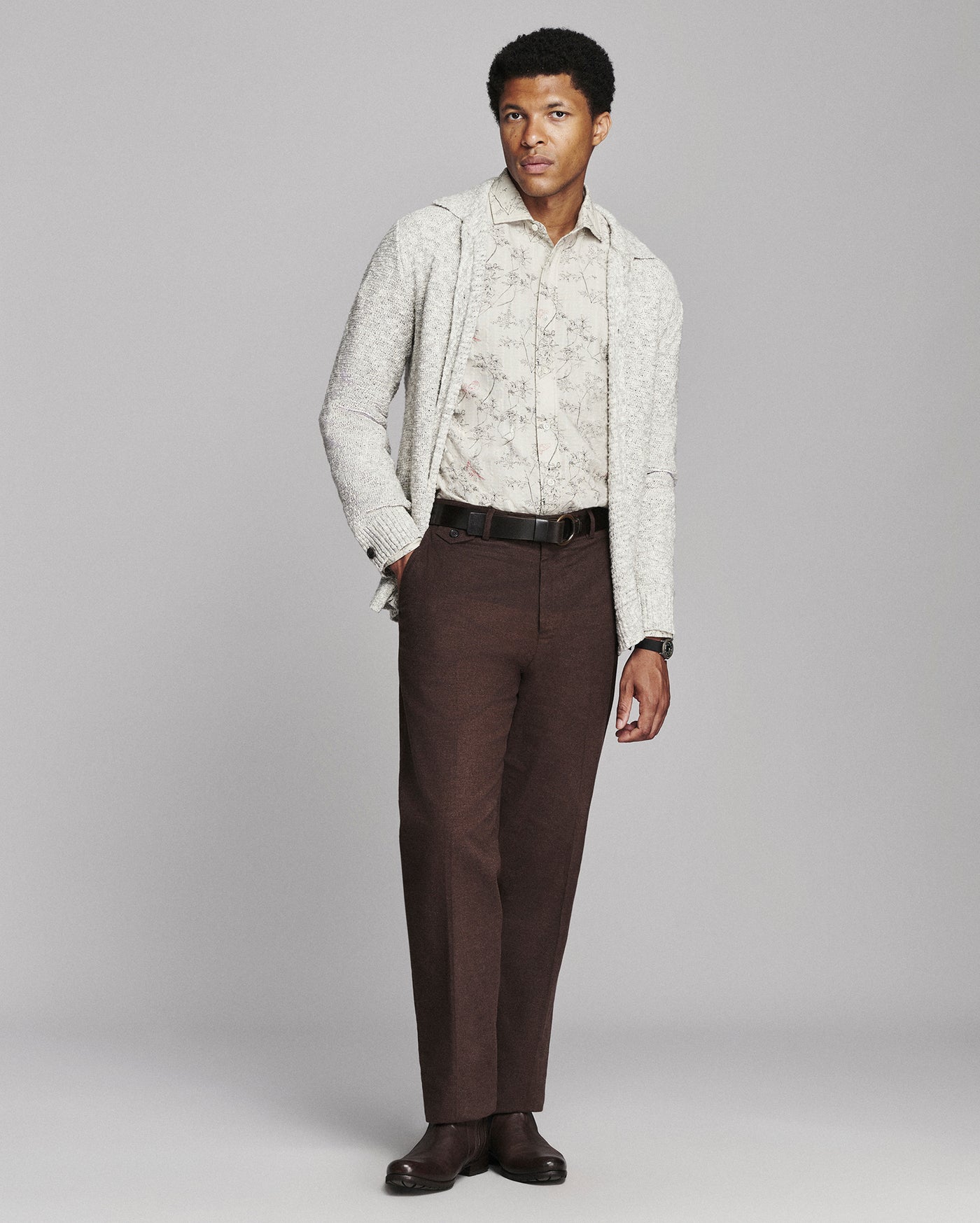 A male model stands against a light gray studio background wearing a cream knit cardigan over a light floral shirt, dark brown trousers, a black belt, and dark brown dress shoes.