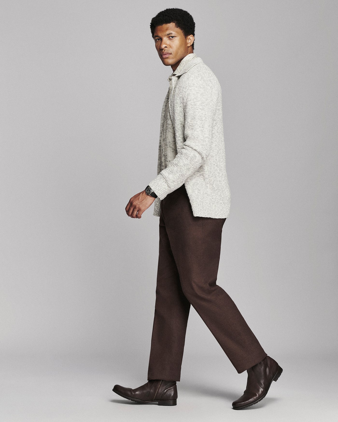 A man in a light gray textured knit cardigan, brown trousers, and dark brown leather boots walking against a solid gray studio background.