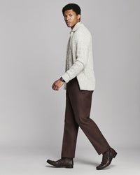 A man in a light gray textured knit cardigan, brown trousers, and dark brown leather boots walking against a solid gray studio background.