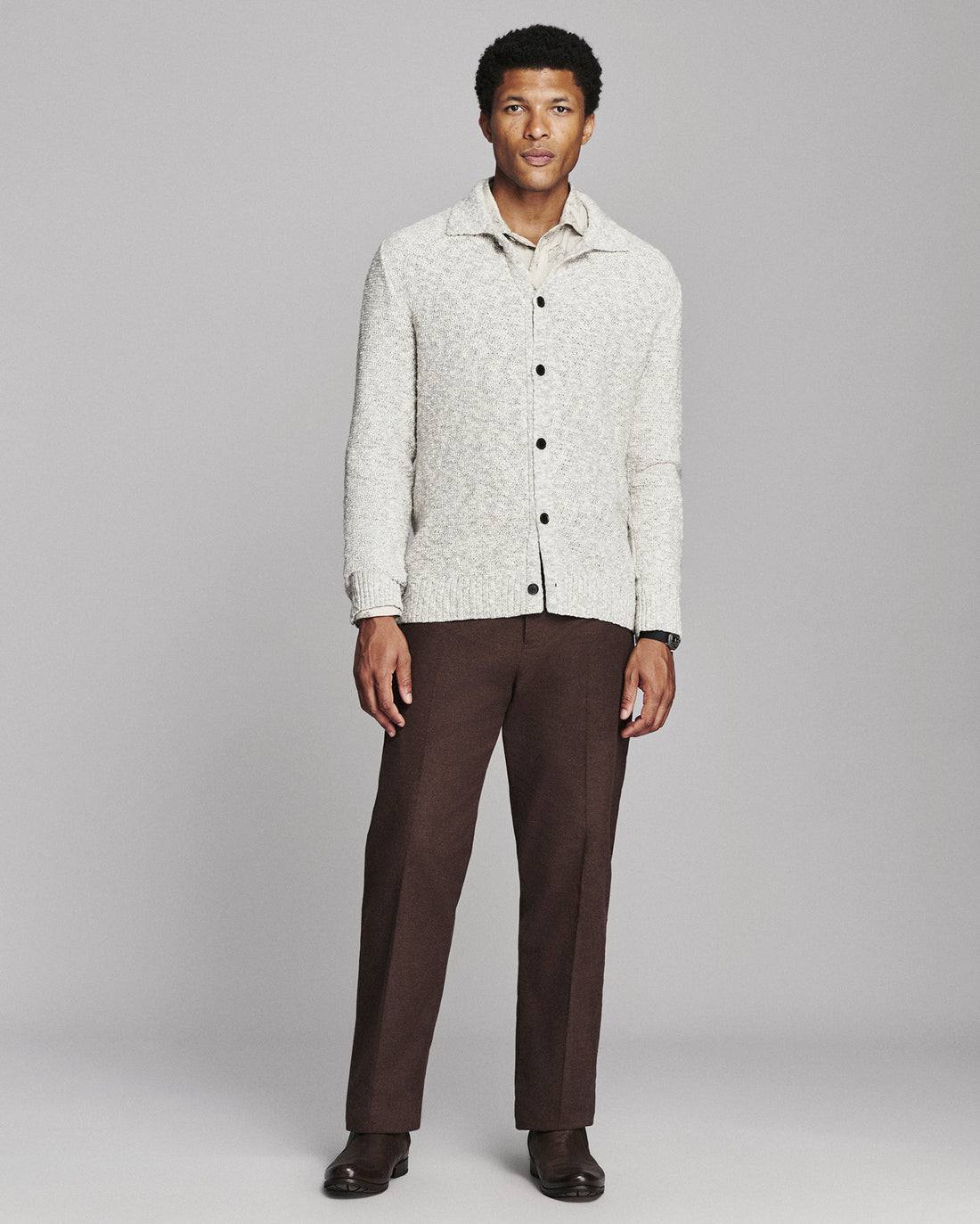 A man modeling a light cream textured knit button-up cardigan over a collared shirt, paired with dark brown trousers and dark brown dress shoes, against a solid gray studio background.