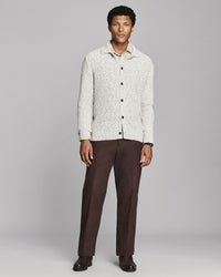 A man modeling a light cream textured knit button-up cardigan over a collared shirt, paired with dark brown trousers and dark brown dress shoes, against a solid gray studio background.
