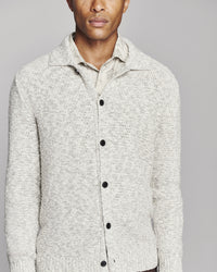 A man wearing a light beige textured knit cardigan sweater with a collar and black buttons, layered over a matching light beige collared shirt, against a solid gray background.