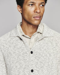 A close-up shot of a man wearing a textured, light gray and white speckled knit cardigan over a beige collared shirt, set against a solid gray studio background.