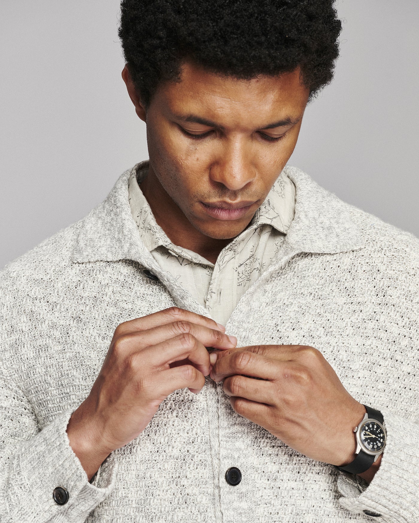 A close-up of a man wearing a textured light gray knit overshirt over a light-colored collared shirt, looking down as he buttons the overshirt. He is wearing a black watch on his wrist.