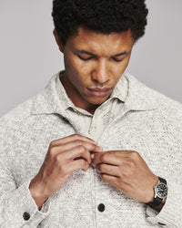 A close-up of a man wearing a textured light gray knit overshirt over a light-colored collared shirt, looking down as he buttons the overshirt. He is wearing a black watch on his wrist.