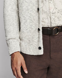 Close-up of a person wearing a light heather gray knit cardigan with dark buttons, a cream patterned shirt, and brown trousers with a black belt.