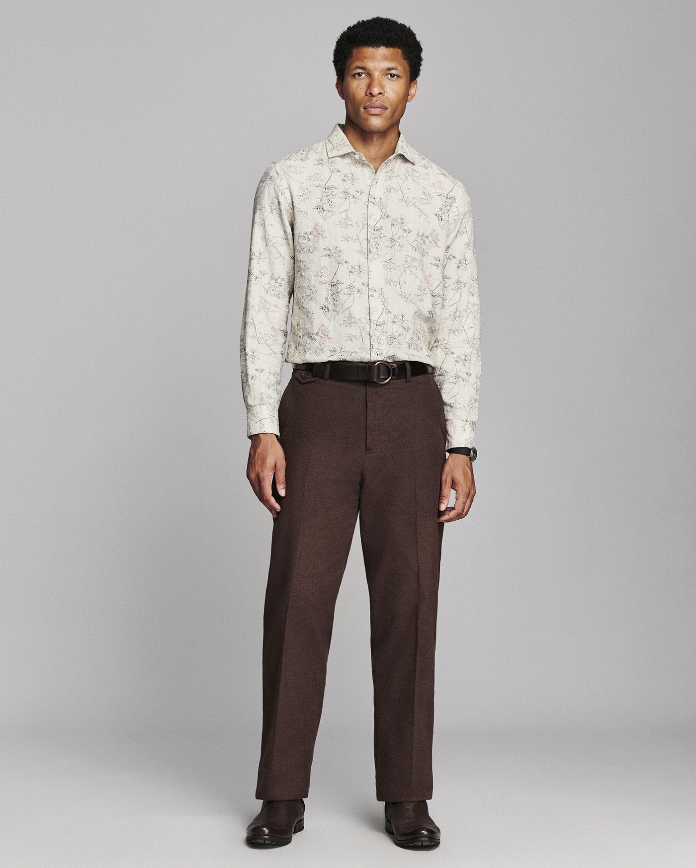 A man modeling a long-sleeve button-up shirt with a light beige and brown abstract pattern, paired with dark brown trousers, a black belt, and dark brown dress shoes, against a solid gray studio background.