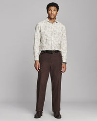 A man modeling a long-sleeve button-up shirt with a light beige and brown abstract pattern, paired with dark brown trousers, a black belt, and dark brown dress shoes, against a solid gray studio background.