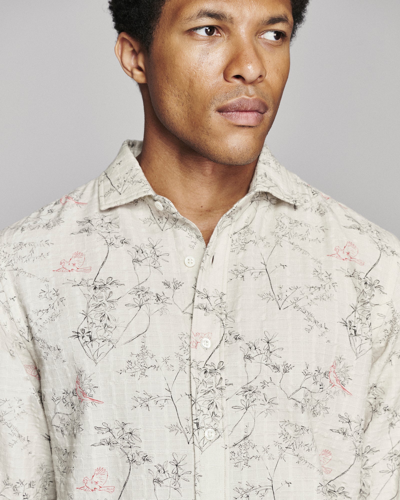 A close-up shot of a man wearing a light beige button-down shirt with a delicate black and pink floral pattern, set against a solid gray background.