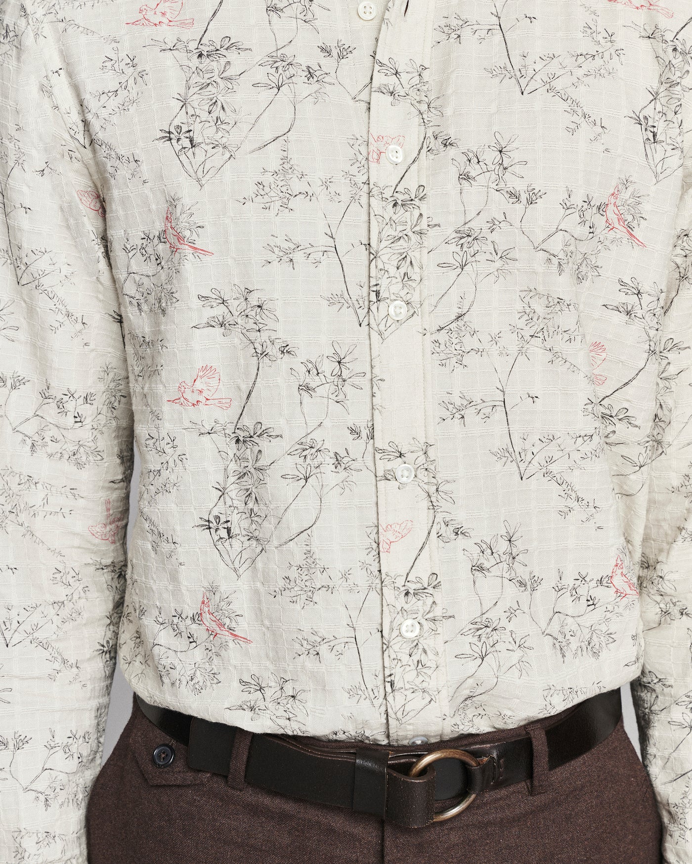 A close-up of a long-sleeve button-up shirt with a delicate botanical and bird print on a light beige fabric, styled with a dark brown belt and dark brown trousers.